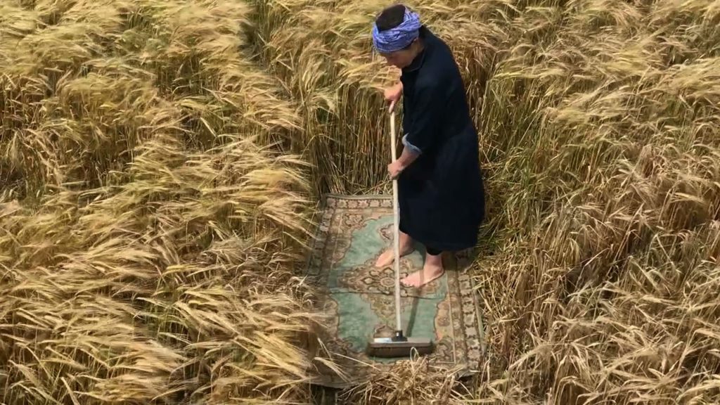 film still of woman sweeping arug in the middle of a field